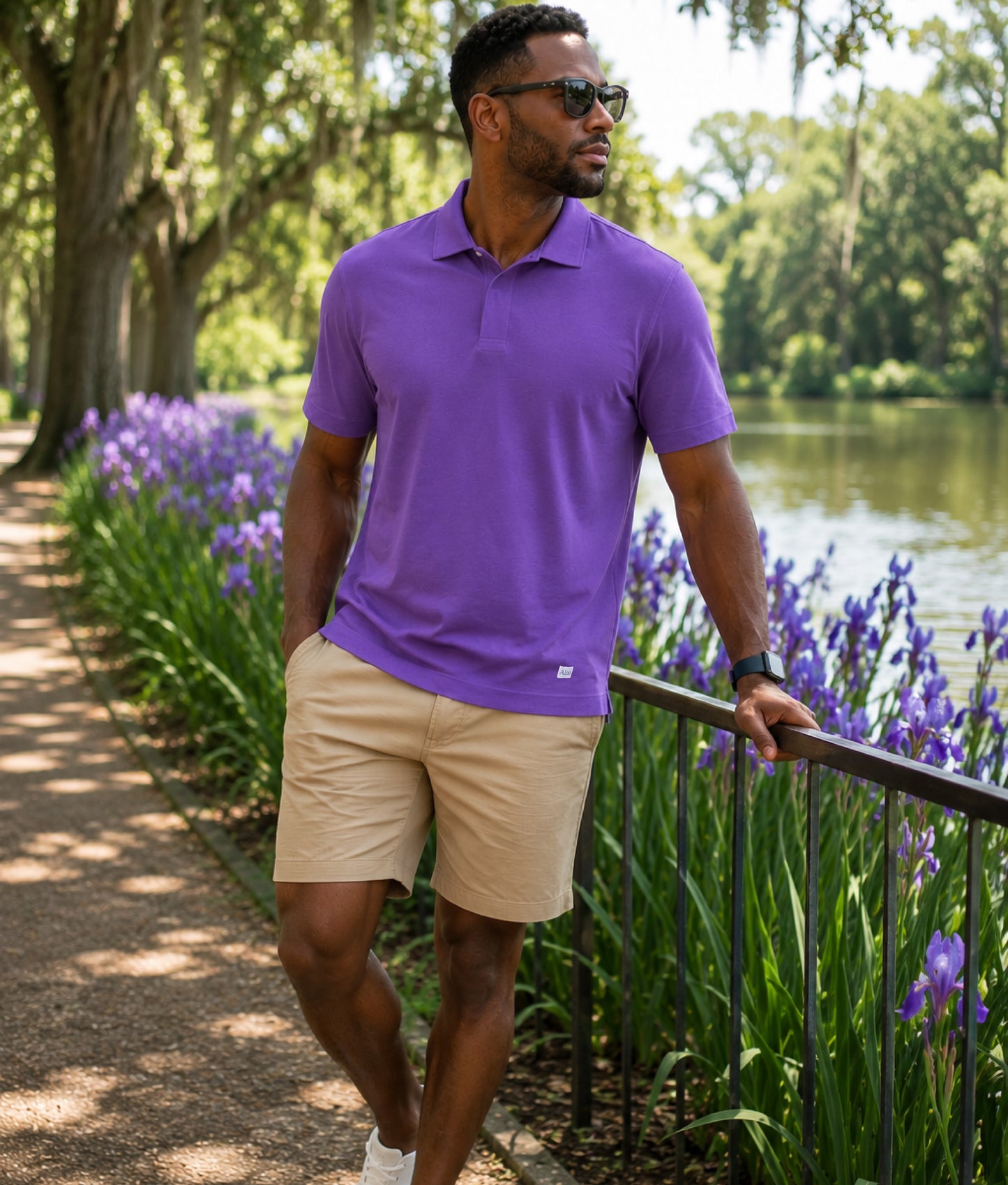 Man in a purple shirt and beige shorts standing by a lake with purple flowers.