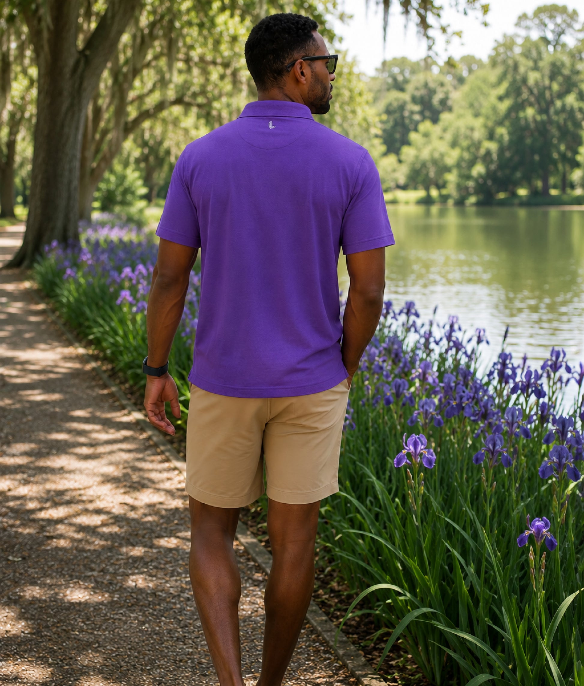Man in a purple shirt and beige shorts walking along a path with purple flowers and a lake in the background.