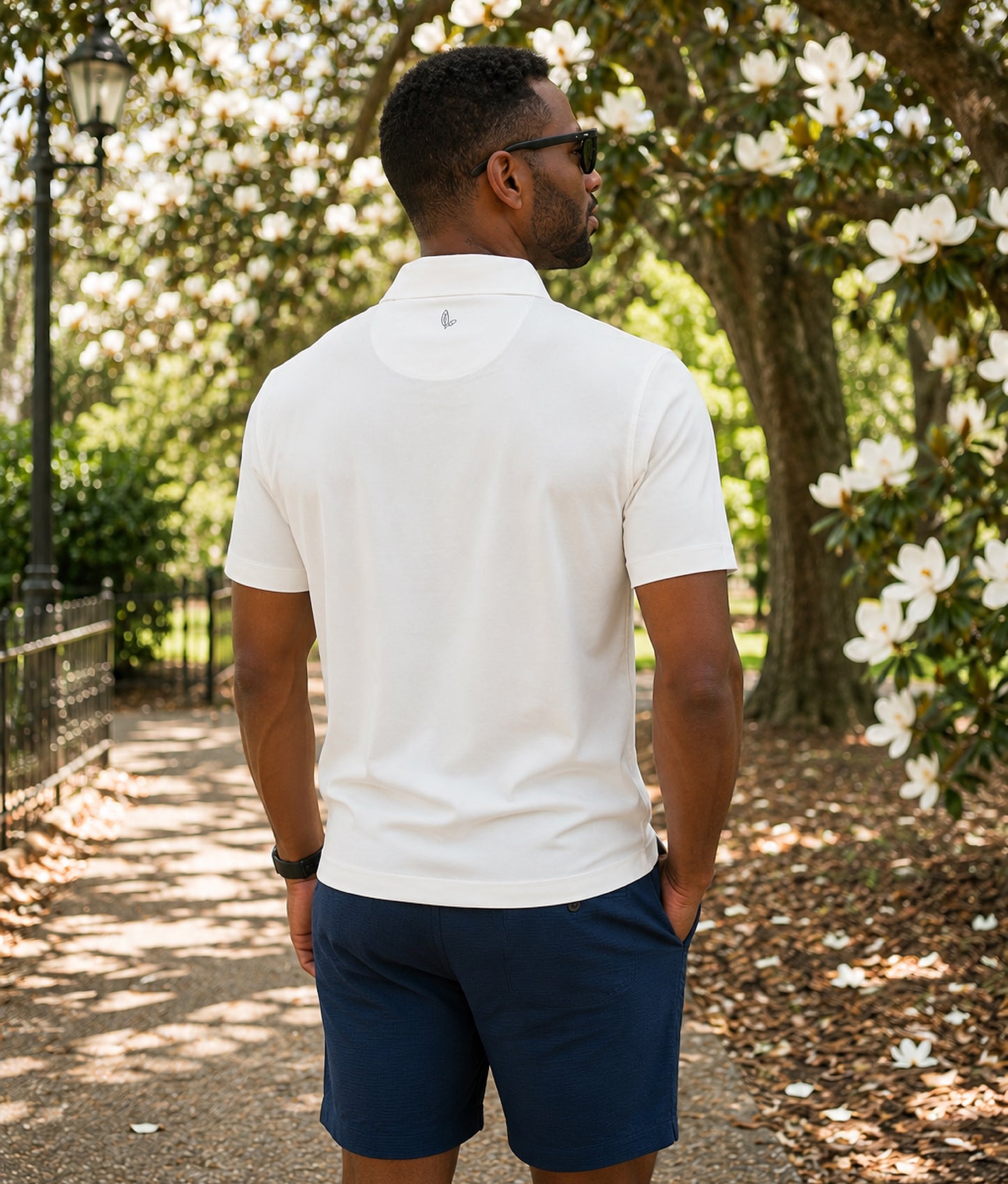 Man wearing a white polo shirt and navy shorts standing in a park with magnolia trees and flowers.