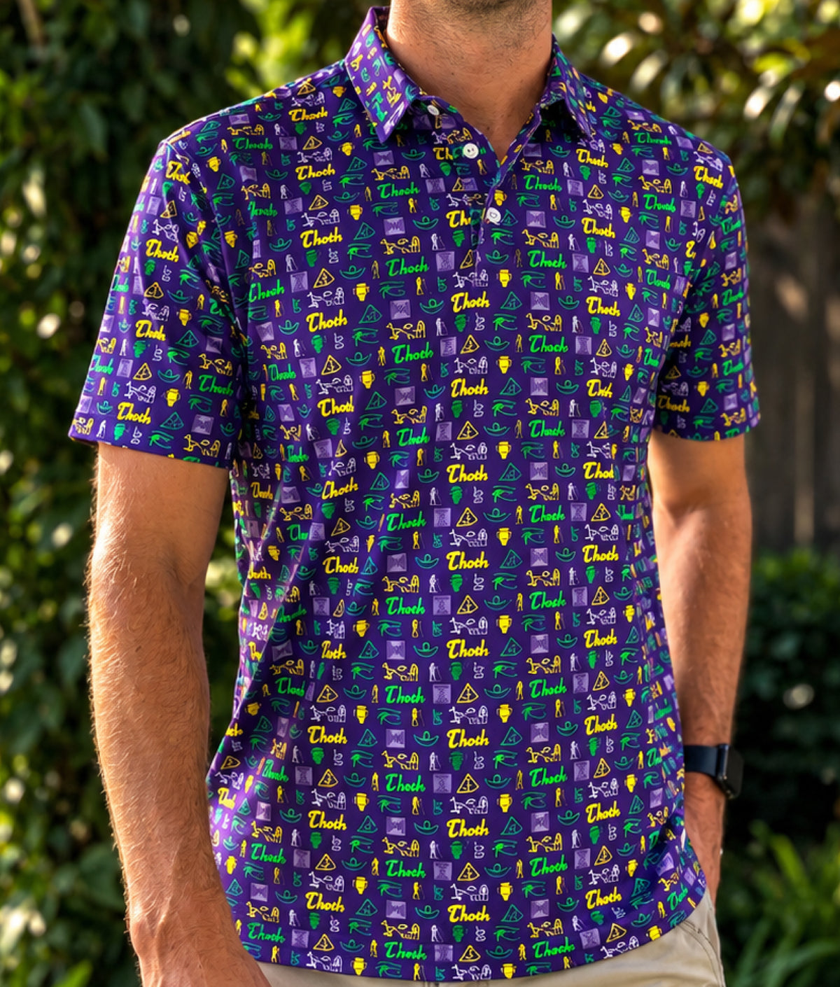 A man wearing a polo shirt with a purple base and an all-over repeating pattern of green, gold, and lavender Thoth/Egyptian themed icons.