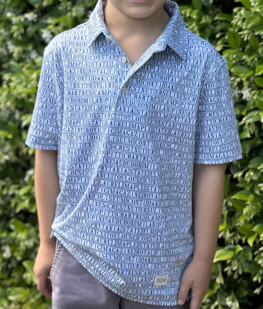 Child wearing a New Orleans street tile patterned polo standing in front of blooming jasmine.