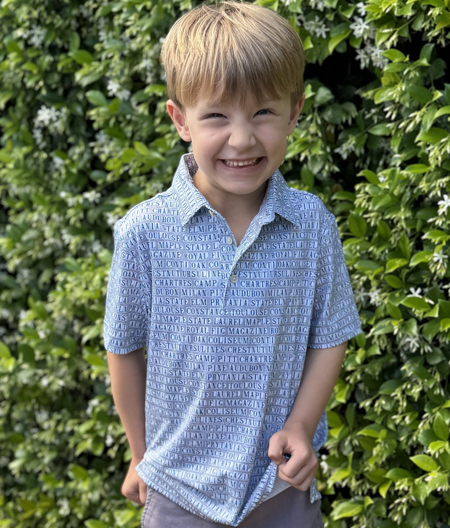 Child wearing a New Orleans street tile patterned polo standing in front of blooming jasmine.